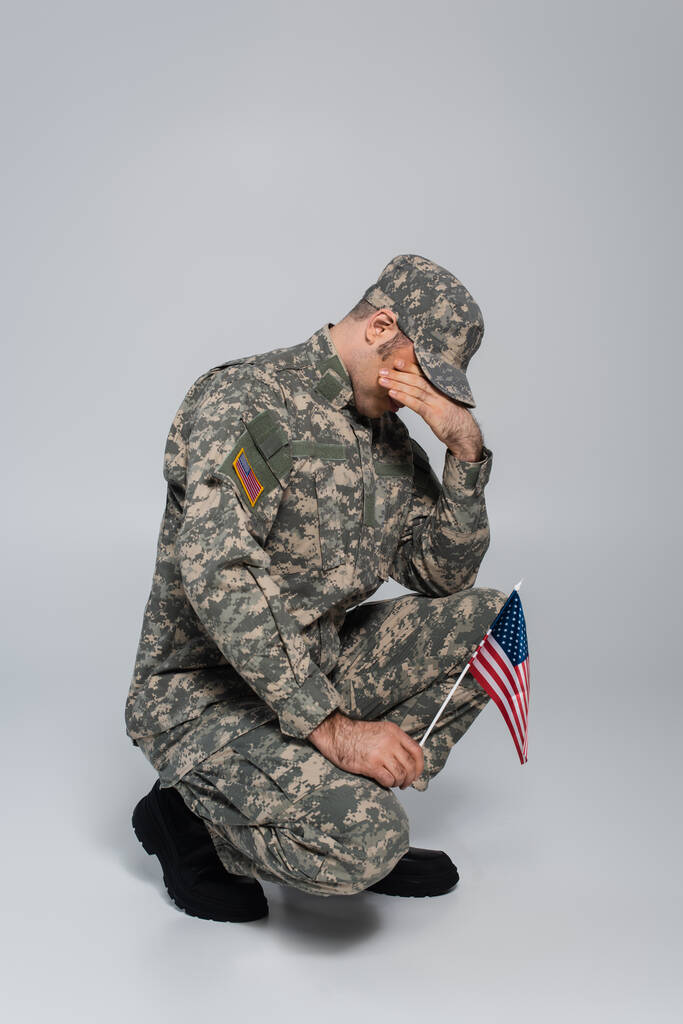 soldado del ejército llorando y cubriendo la cara mientras sostiene la bandera de Estados Unidos durante el día conmemorativo en gris  - Foto, imagen