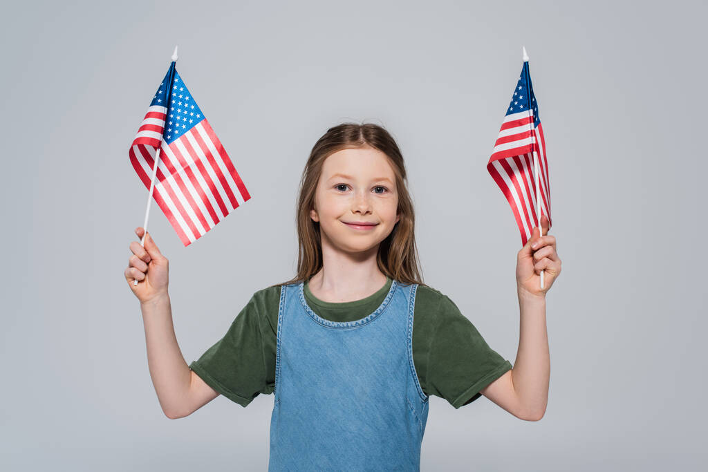 patriotic preteen girl smiling while holding flags of America during memorial day isolated on grey  - Photo, Image
