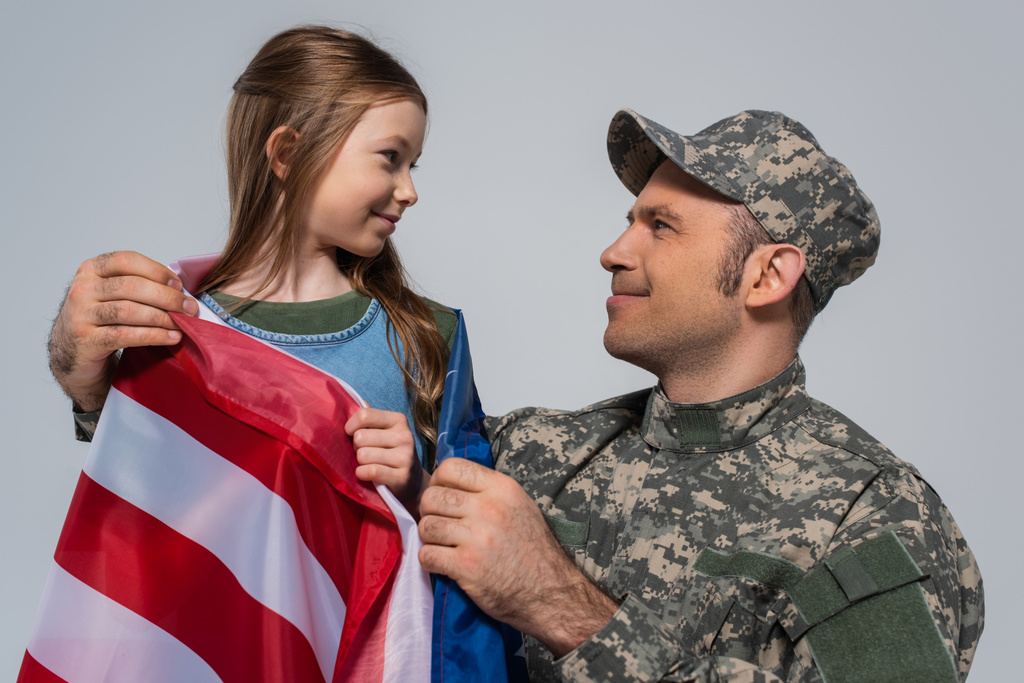 στρατιώτης με στρατιωτική στολή κοιτάζοντας ευτυχισμένη κόρη τυλιγμένη με σημαία των Ηνωμένων Πολιτειών της Αμερικής απομονωμένη σε γκρι  - Φωτογραφία, εικόνα