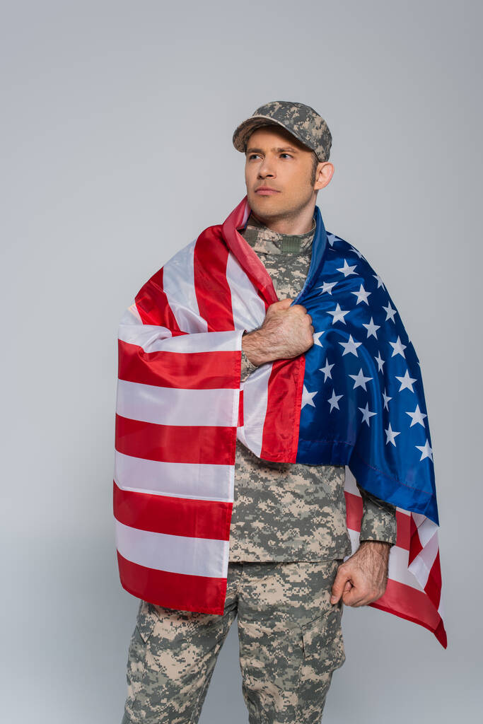 Patriotischer Armeesoldat in Tarnuniform, eingehüllt in die Flagge der Vereinigten Staaten von Amerika am Gedenktag isoliert auf grau  - Foto, Bild
