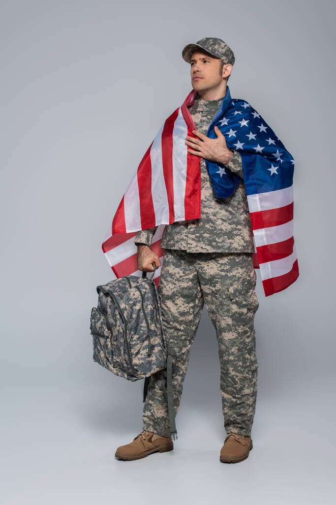 patriottische leger soldaat in camouflage uniform verpakt in vlag van de Verenigde Staten van Amerika staan met rugzak op grijs  - Foto, afbeelding