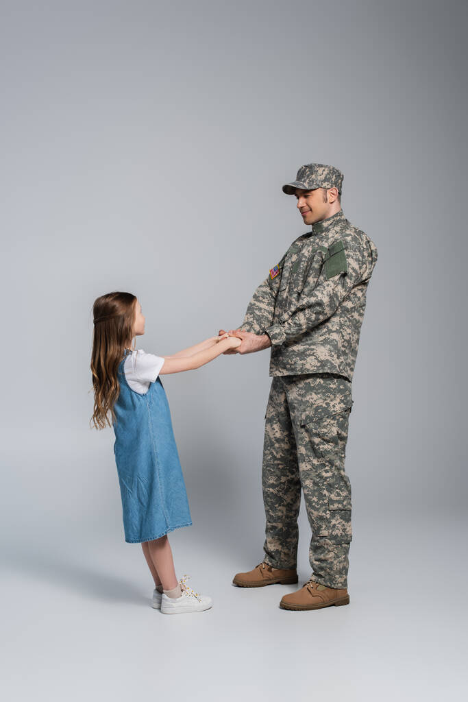 pełna długość szczęśliwy żołnierz w mundurze wojskowym trzymając się za ręce z córką na szarości  - Zdjęcie, obraz