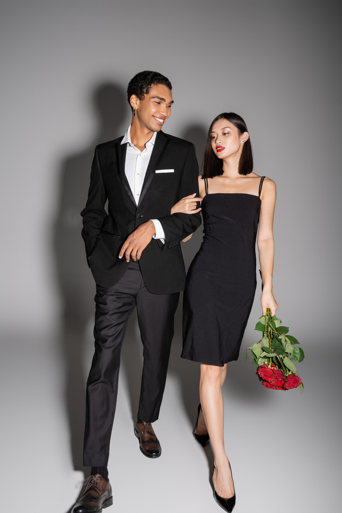 longitud completa de hombre afroamericano feliz y elegante mujer asiática con rosas rojas caminando sobre fondo gris - Foto, imagen