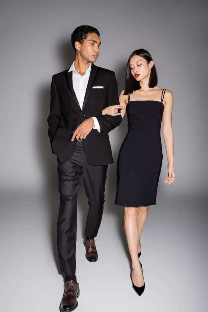 volle Länge des multiethnischen Paares in schwarzer eleganter Kleidung, das auf grauem Hintergrund wandelt - Foto, Bild