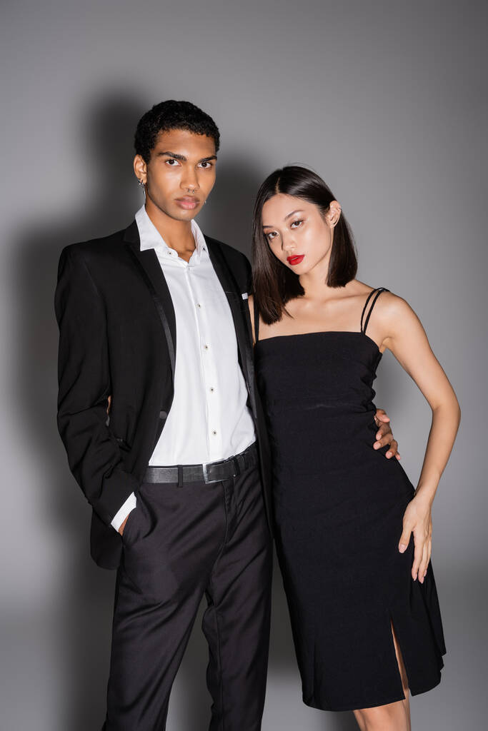 african american man in black suit and asian woman in strap dress looking at camera on grey background - Photo, Image