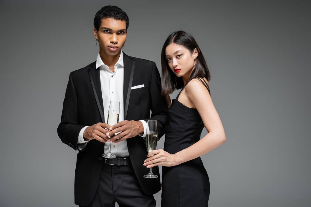 афроамериканський чоловік в чорному костюмі і азіатська жінка в сукні ремінця тримає шампанське і дивиться на камеру ізольовано на сірому
 - Фото, зображення