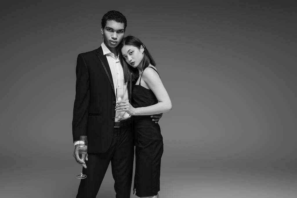 giovane coppia interrazziale in elegante abbigliamento formale in posa con bicchieri di champagne isolati su grigio - Foto, immagini