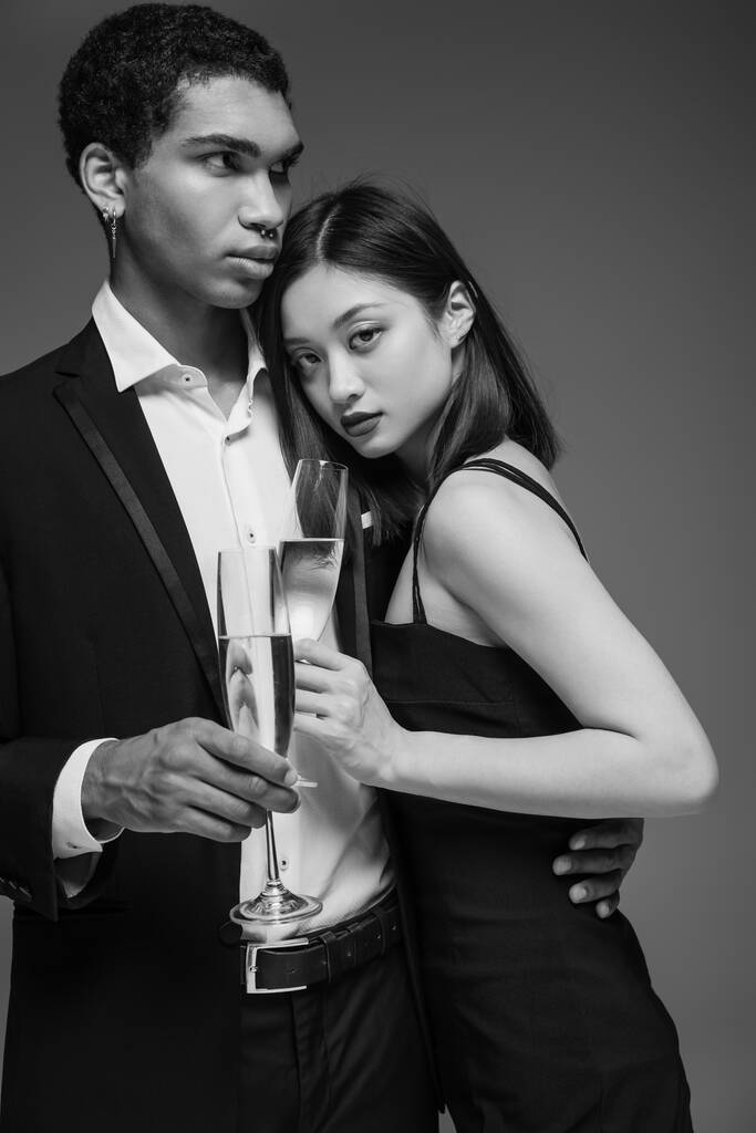 pareja interracial joven y de moda en ropa formal sosteniendo copas de champán aisladas en gris - Foto, Imagen
