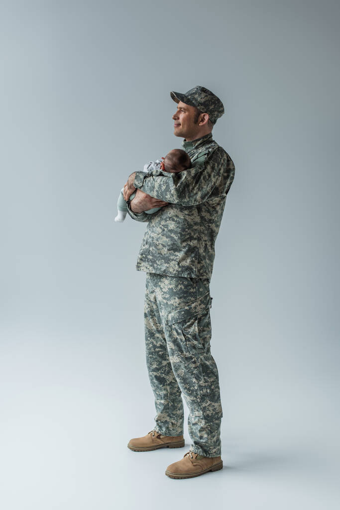повна довжина приємного військовослужбовця в формі і кепка тримає новонародженого сина на сірому
  - Фото, зображення