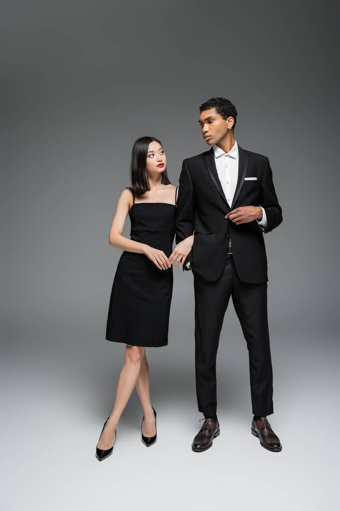 longitud completa de pareja interracial en traje elegante negro mirándose el uno al otro sobre fondo gris - Foto, imagen