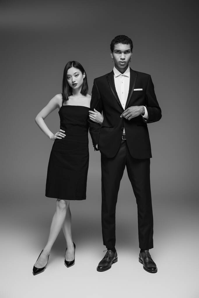 teljes hossza afro-amerikai férfi fekete öltöny és ázsiai nő elegáns ruha pózol szürke háttér - Fotó, kép