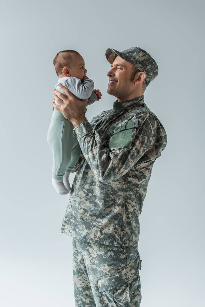 soldado militar feliz em uniforme segurando filho recém-nascido em armas isoladas em cinza  - Foto, Imagem