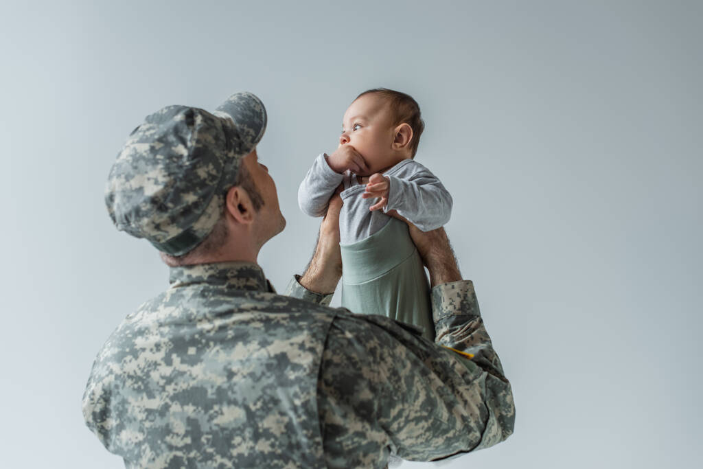 leger soldaat in uniform en pet houden baby jongen in armen geïsoleerd op grijs  - Foto, afbeelding