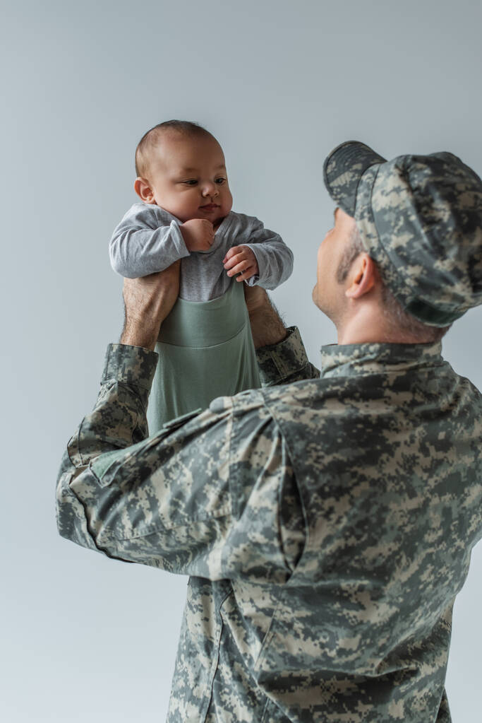 soldado do exército em uniforme militar com boné segurando filho bebê em armas isoladas em cinza - Foto, Imagem