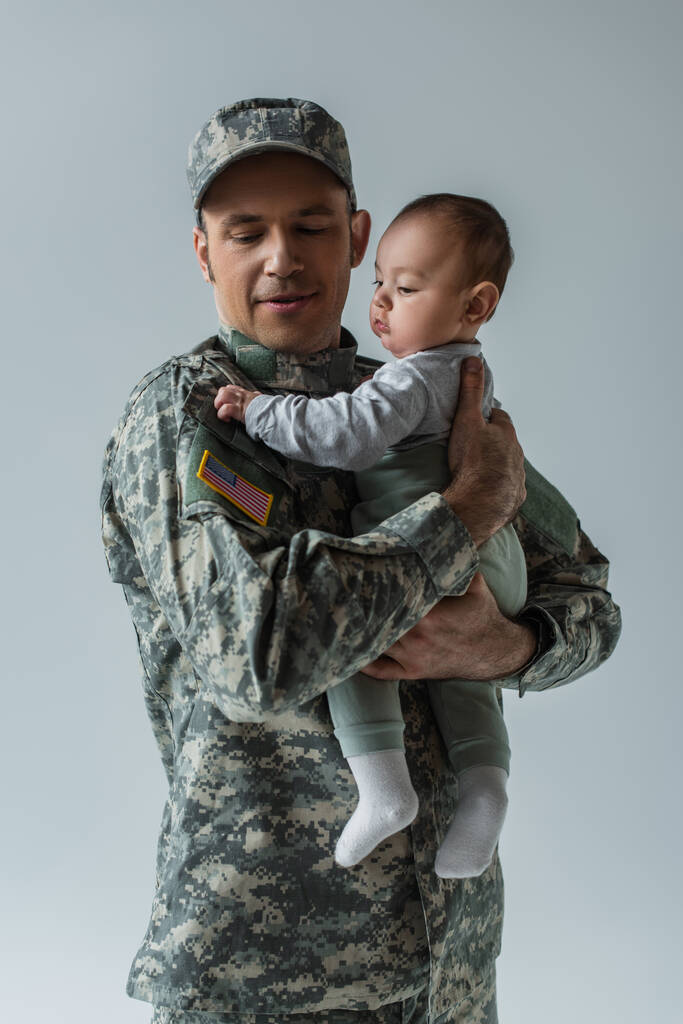 Amerikaanse leger soldaat in militair uniform met pet houdt zuigeling zoon in armen geïsoleerd op grijs - Foto, afbeelding
