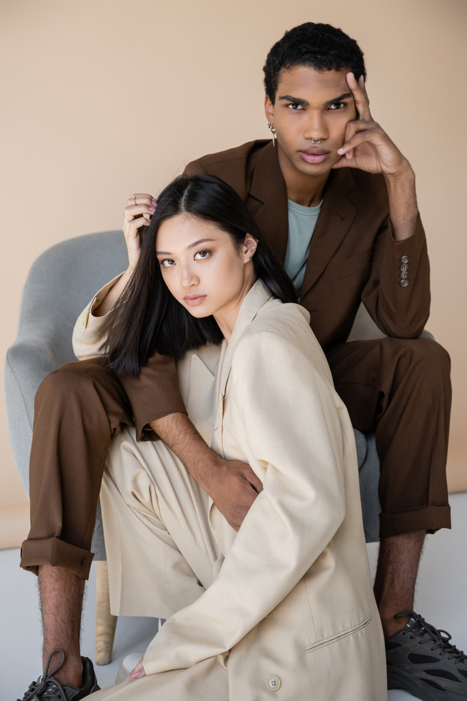 modieuze interraciale modellen in trendy broekpakken kijken naar camera in de buurt van fauteuil op beige achtergrond - Foto, afbeelding