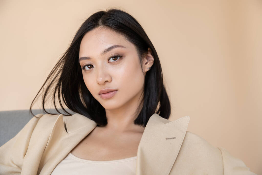 portrait of brunette asian woman in ivory blazer looking at camera on beige background - Photo, Image