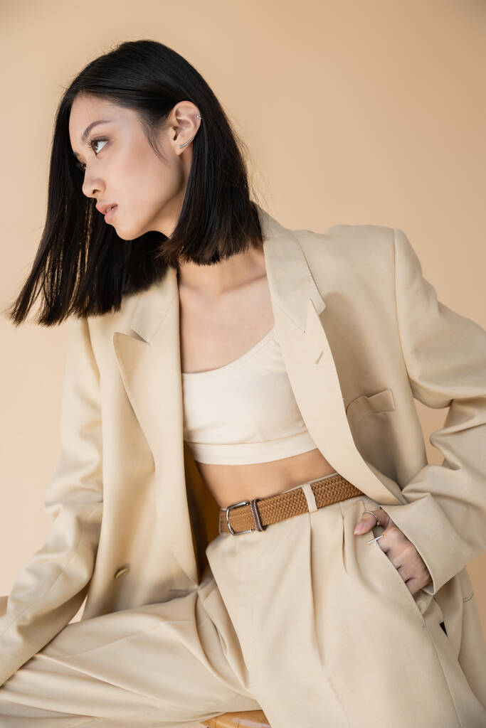 brunette asian woman in stylish pantsuit posing with hand in pocket and looking away on beige background - Photo, Image