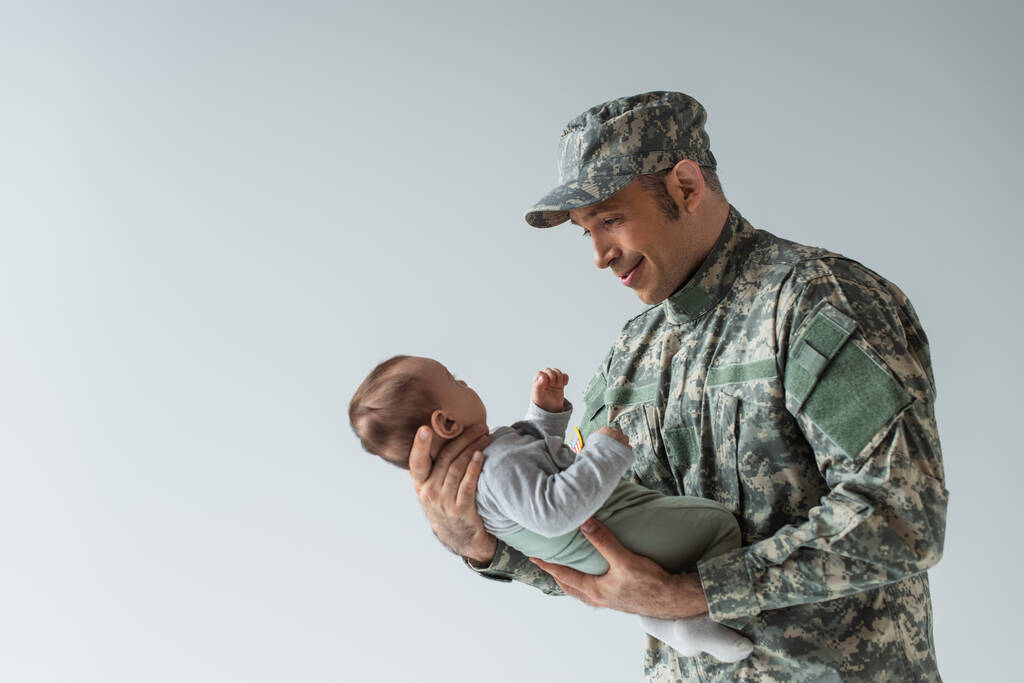 Üniformalı mutlu Amerikan askeri yeni doğan çocuğa bakıyor ve gri renkte sırıtıyor.  - Fotoğraf, Görsel
