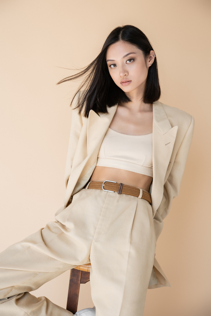 alla moda donna asiatica in pantalone posa vicino sgabello e guardando la fotocamera isolata sul beige - Foto, immagini