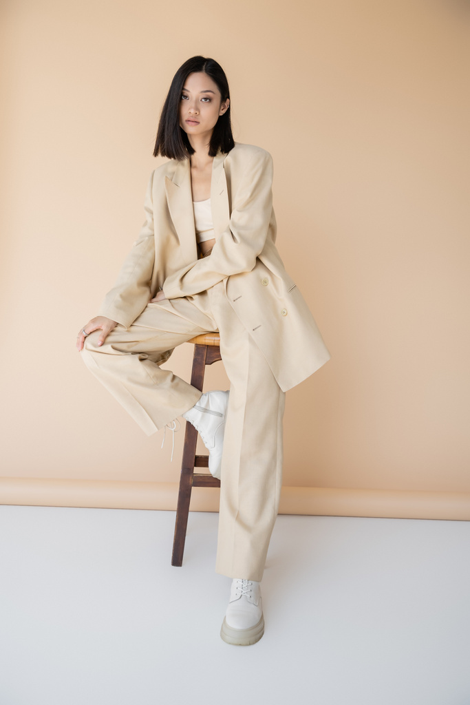 brunette aasialainen nainen tyylikäs housupuku ja saappaat istuu jakkaralla beige tausta - Valokuva, kuva