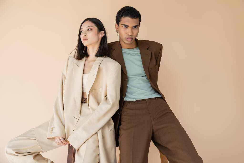 homme afro-américain à la mode en costume marron regardant la caméra près de brunette modèle asiatique isolé sur beige - Photo, image