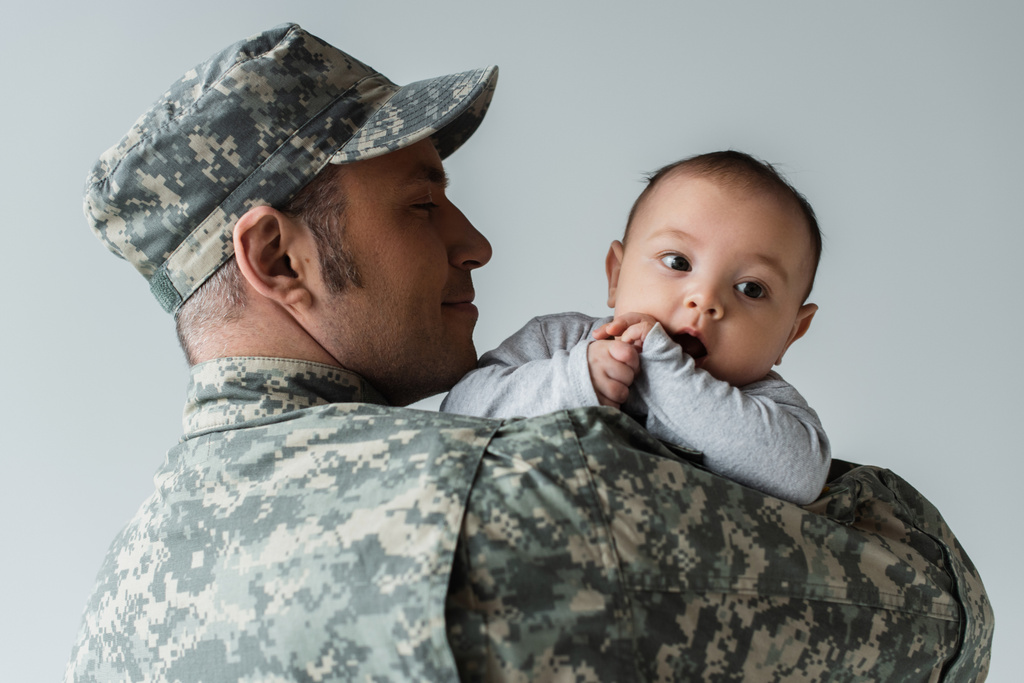 Χαρούμενος πατέρας με στρατιωτική στολή και καπέλο αγκαλιάζει νεογέννητο γιο απομονωμένο στο γκρι  - Φωτογραφία, εικόνα