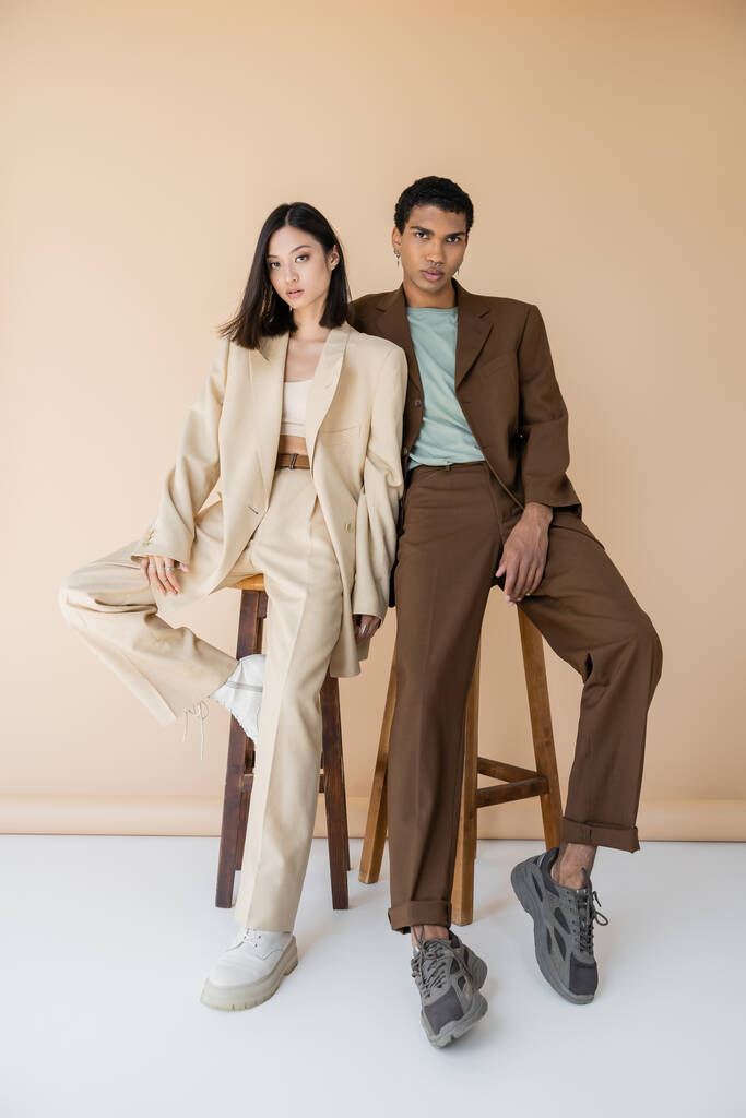 comprimento total de casal multiétnico em pantsuits elegantes posando perto de fezes no fundo bege - Foto, Imagem