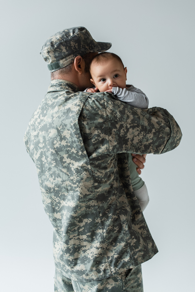 человек в военной форме и военном колпаке обнимает новорожденного сына, изолированного на сером  - Фото, изображение