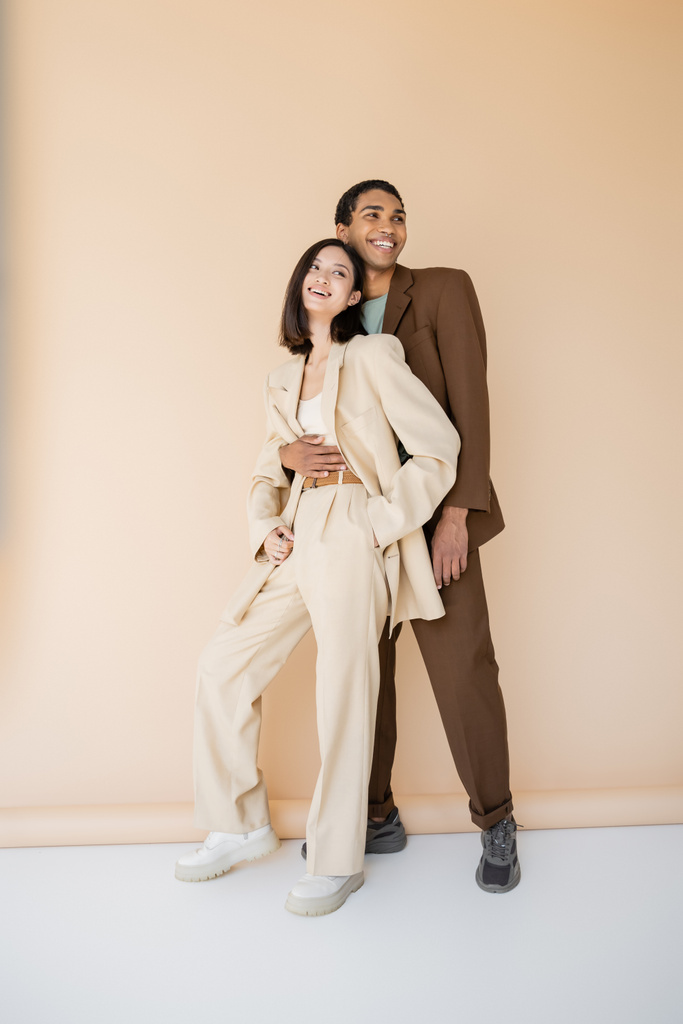 comprimento total de homem americano africano feliz em terno marrom abraçando mulher asiática elegante no fundo bege - Foto, Imagem