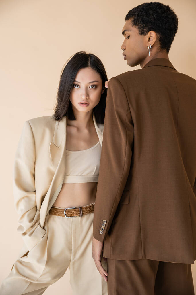 tendance asiatique femme en ivoire pantalon debout avec la main dans la poche près de l'homme afro-américain en blazer brun isolé sur beige - Photo, image