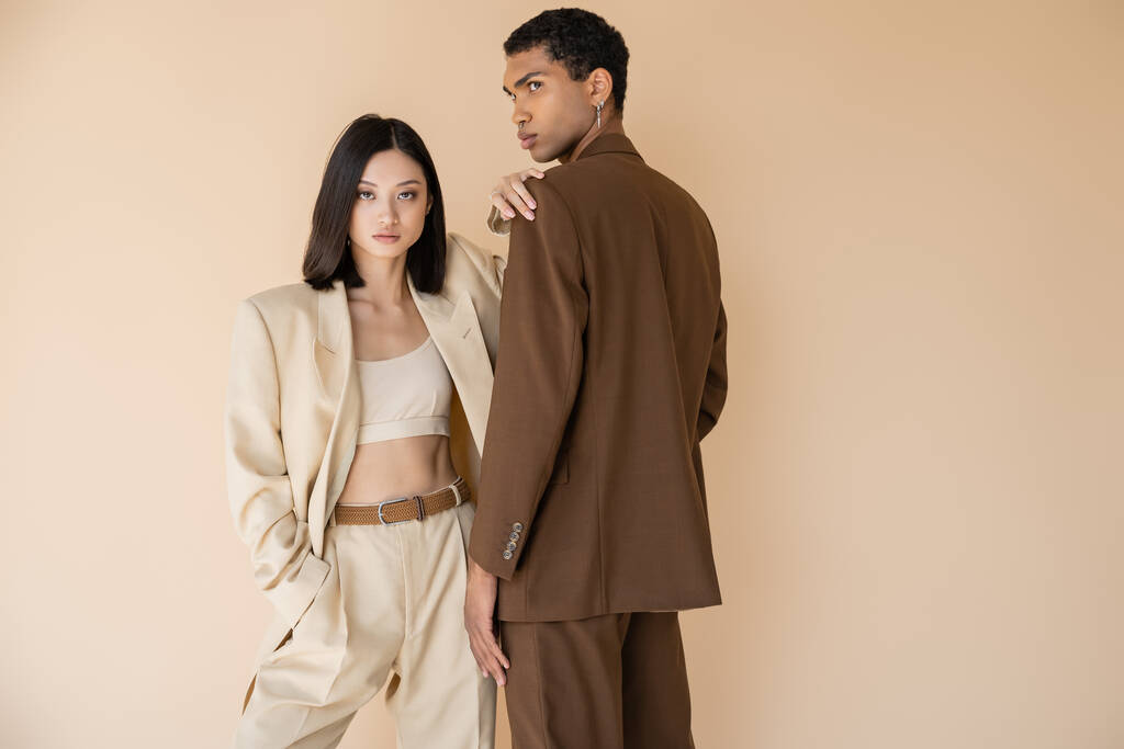 ázsiai nő divatos nadrágkosztümben nézi a kamerát, miközben pózol a zsebében közel afro-amerikai férfi elszigetelt bézs - Fotó, kép