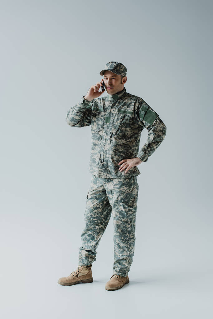 plná délka vojáka v čepici a armádní uniformě mluví na smartphone a stojí na šedé  - Fotografie, Obrázek