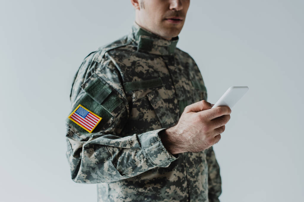 oříznutý pohled na amerického vojáka pomocí chytrého telefonu izolovaného na šedé  - Fotografie, Obrázek