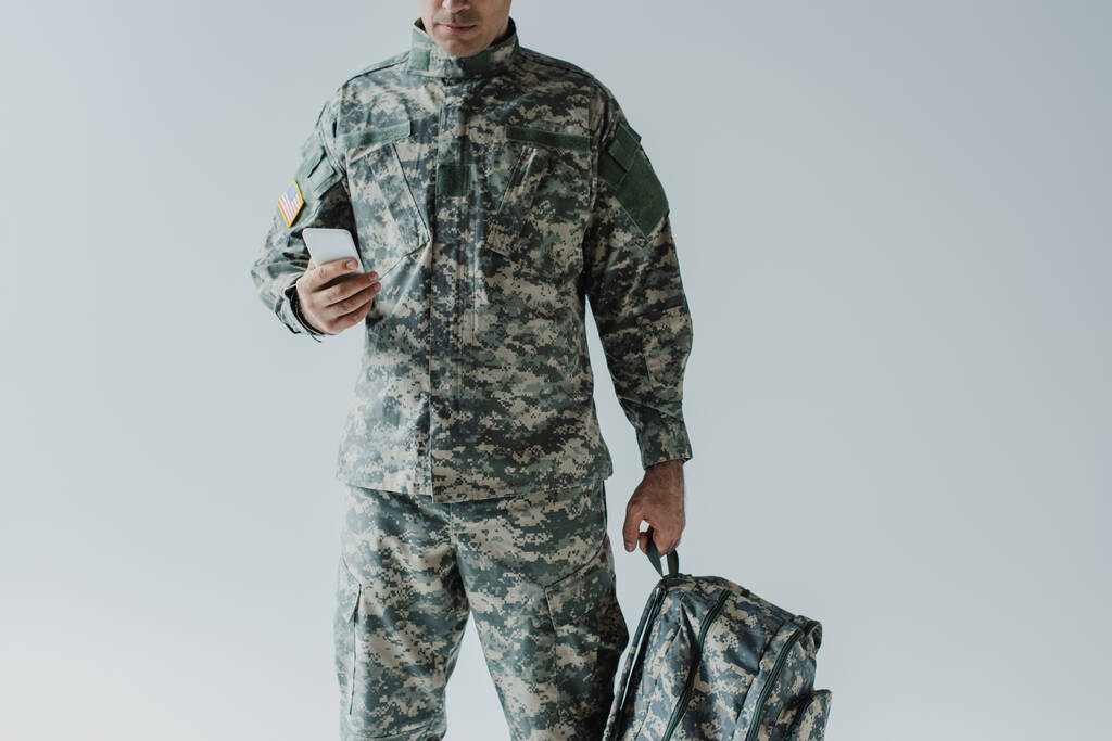 обрезанный вид американского военнослужащего с помощью смартфона во время проведения рюкзак изолирован на серый  - Фото, изображение