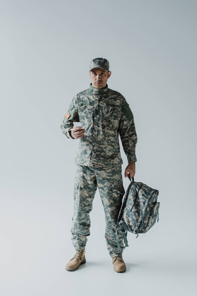 Soldado americano usando teléfono inteligente mientras sostiene la mochila y de pie en gris  - Foto, Imagen