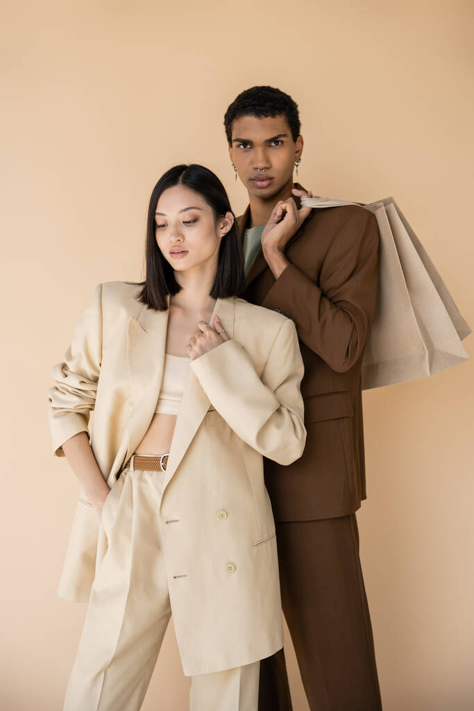 Afrikanisch-amerikanischer Mann mit Einkaufstaschen blickt in die Kamera nahe asiatische Frau in Strumpfhosen isoliert auf beige - Foto, Bild