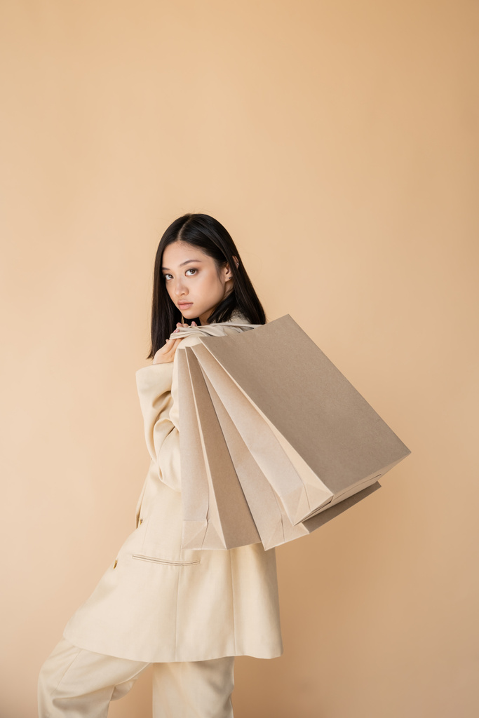 молодая и модная азиатка с сумками, смотрящая на камеру, изолированную от бежевого - Фото, изображение