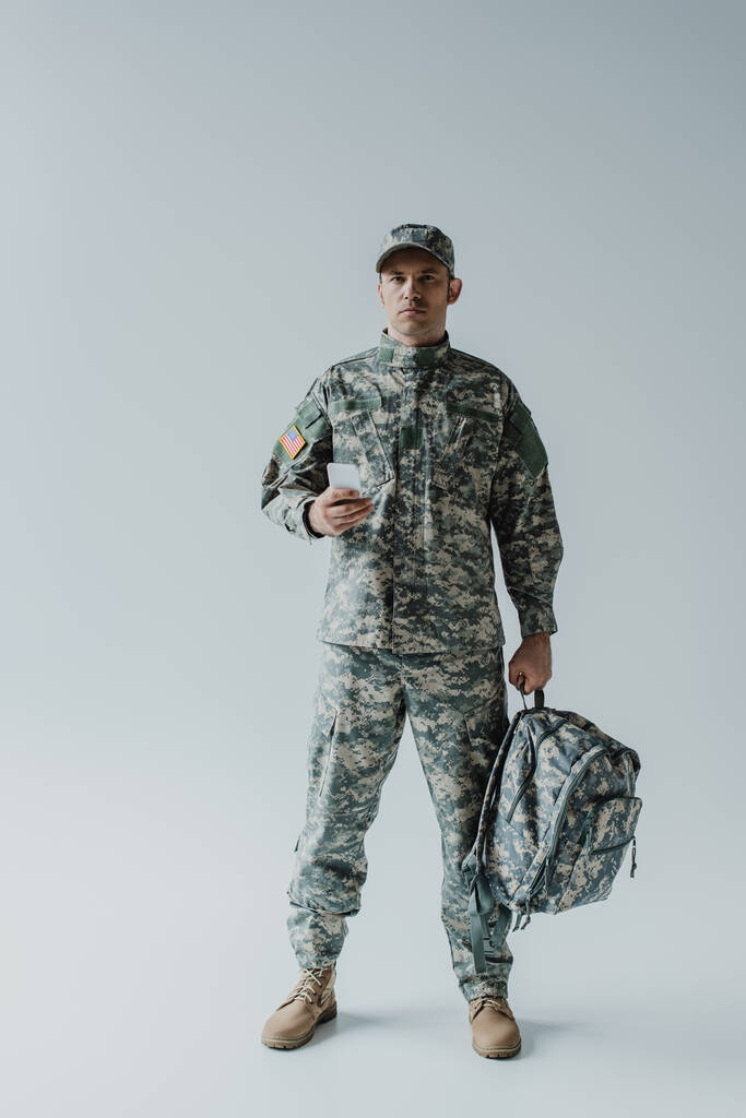 Soldato americano in uniforme militare con bandiera nazionale con smartphone e zaino su grigio  - Foto, immagini