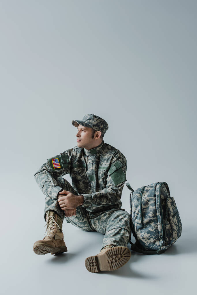 Amerikanischer Soldat in Armeeuniform mit Nationalflagge sitzt neben Rucksack auf grau  - Foto, Bild