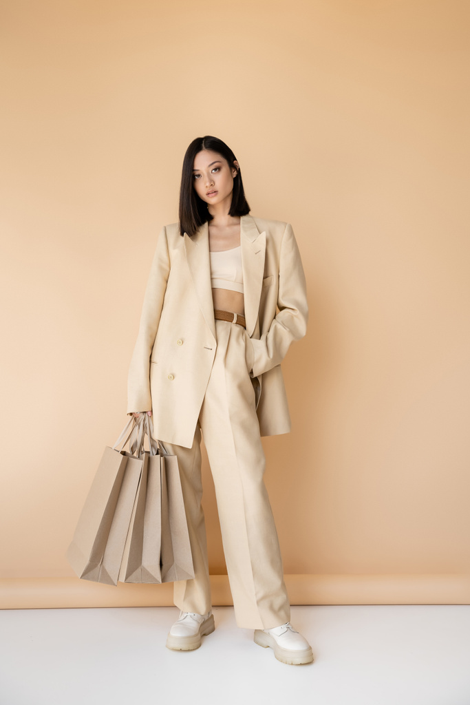 pełna długość brunetka Azji kobieta w stylowy pantsuit stojący z zakupy torby na beżowym tle - Zdjęcie, obraz