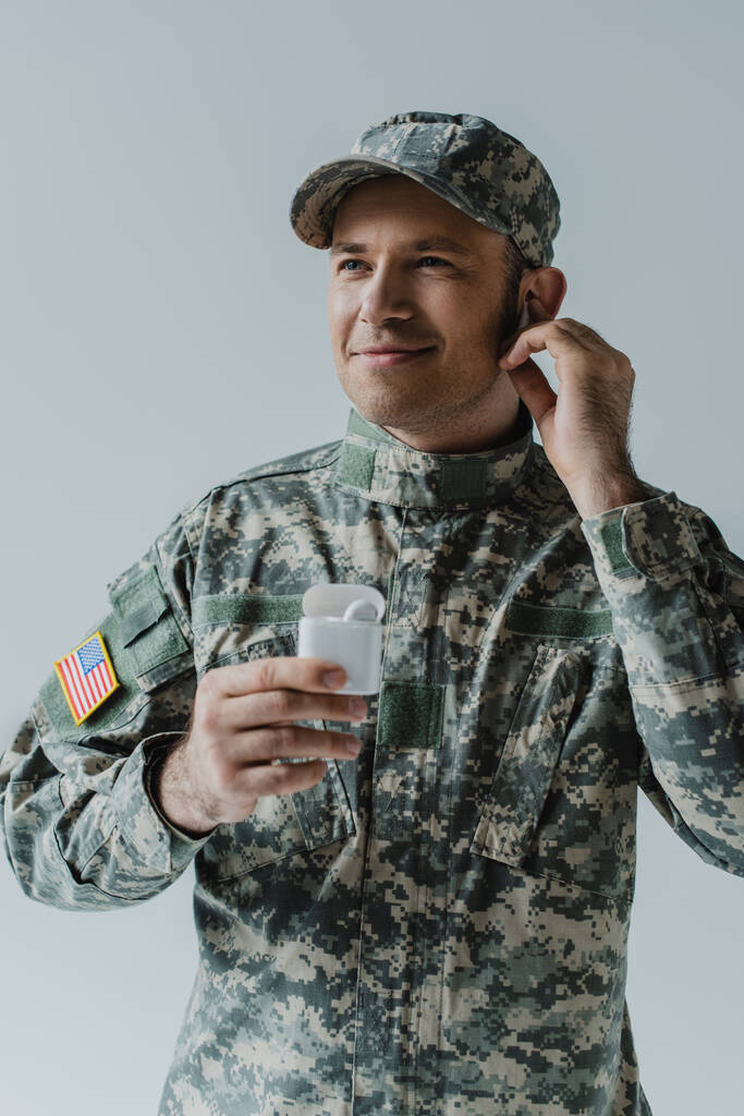 vidám katona egyenruhában és sapkában fülhallgatót tartva és mosolyogva elszigetelve a szürkén.  - Fotó, kép