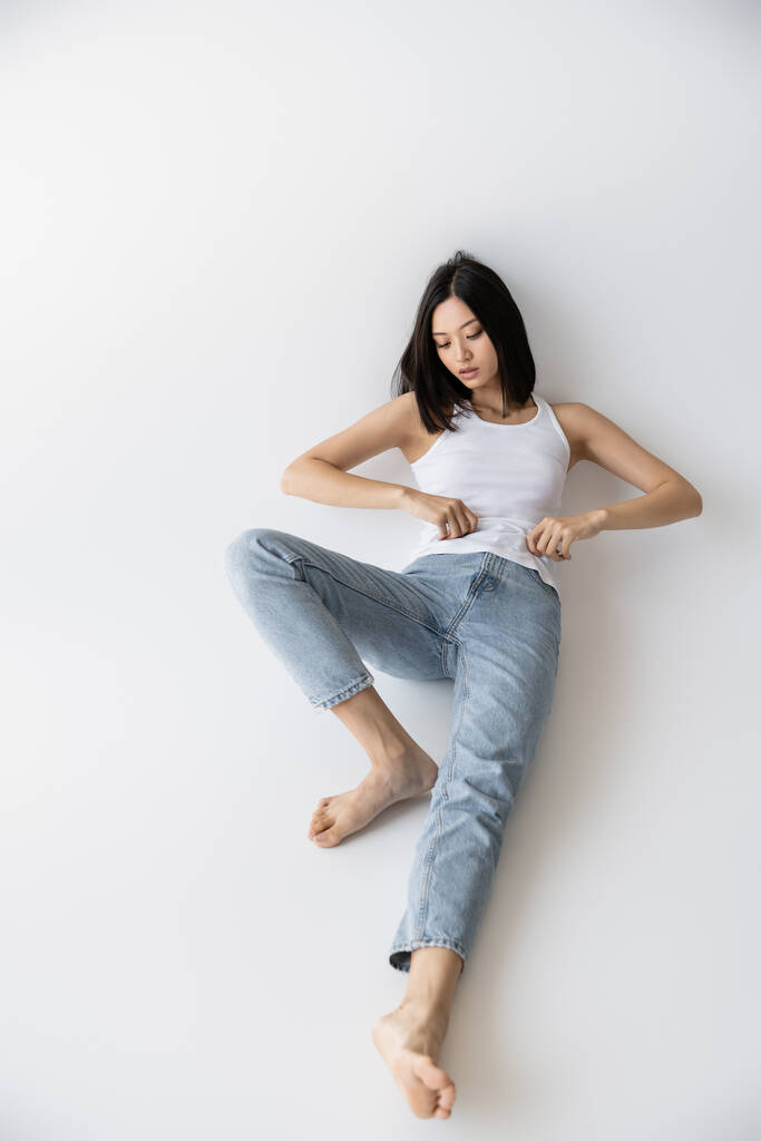 pleine longueur de pieds nus asiatique femme en blanc débardeur et jeans couché sur fond gris - Photo, image