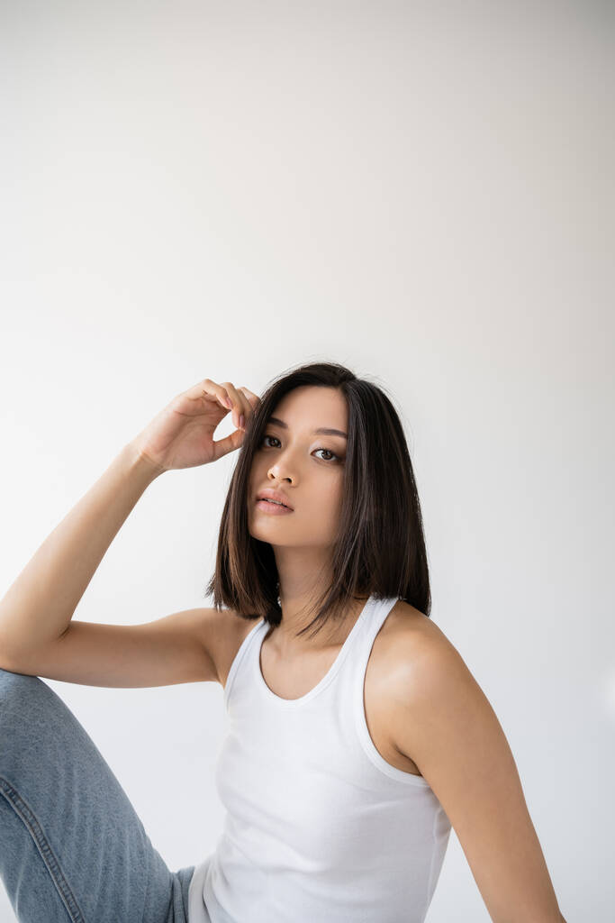 brunette asian woman in white tank top holding hand near face and looking at camera on grey background - Photo, Image