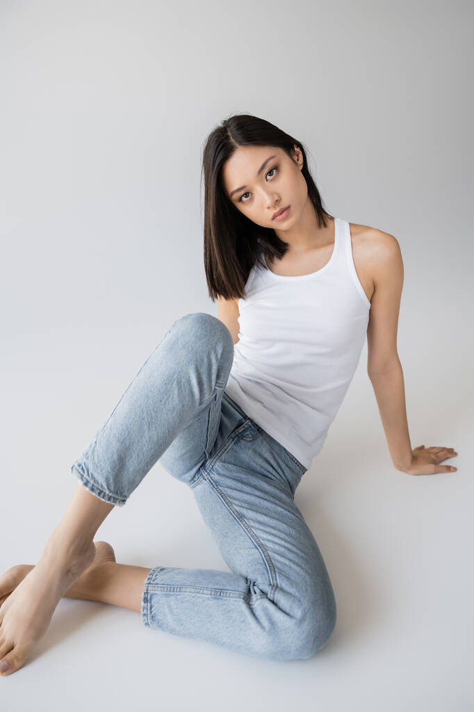 красивая азиатка в белой майке и джинсах, смотрящая на камеру, сидя на сером фоне - Фото, изображение
