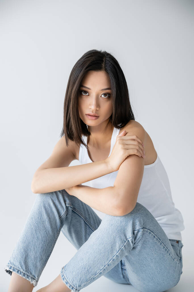 charming asian woman in blue jeans looking at camera while sitting on grey background - Photo, Image