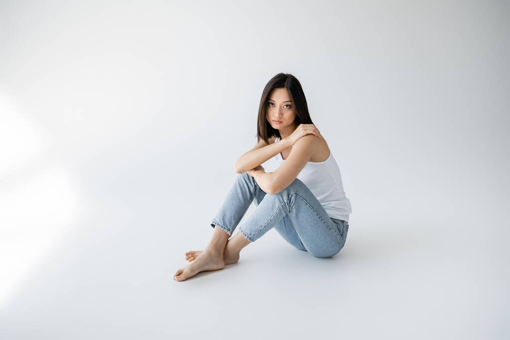 volle Länge der asiatischen Frau in blauen Jeans und Tank-Top schaut in die Kamera, während sie auf grauem Hintergrund sitzt - Foto, Bild