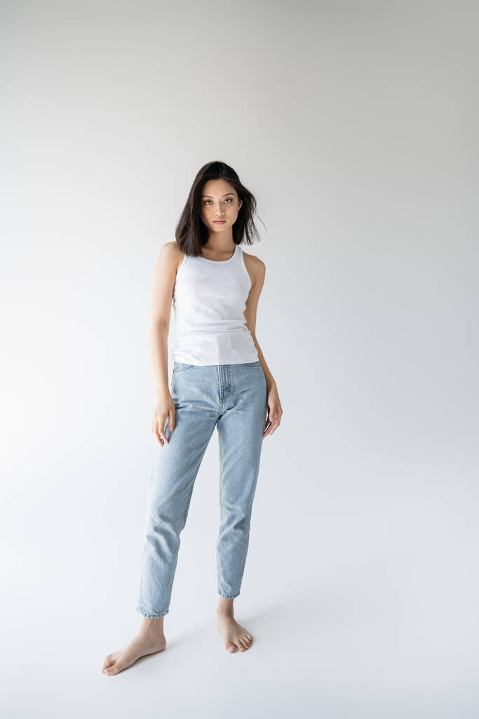 volle Länge der barfuß und schlanke asiatische Frau in Tank Top und Jeans stehen auf grauem Hintergrund - Foto, Bild
