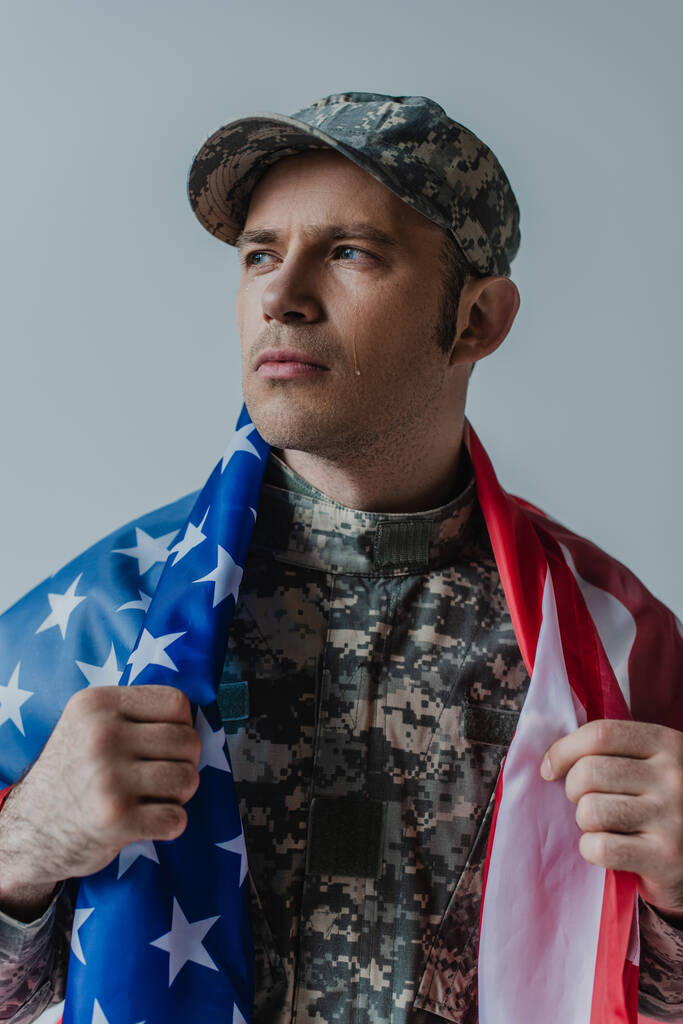 triste soldado americano sosteniendo la bandera de Estados Unidos mientras llora durante el día conmemorativo aislado en gris  - Foto, Imagen