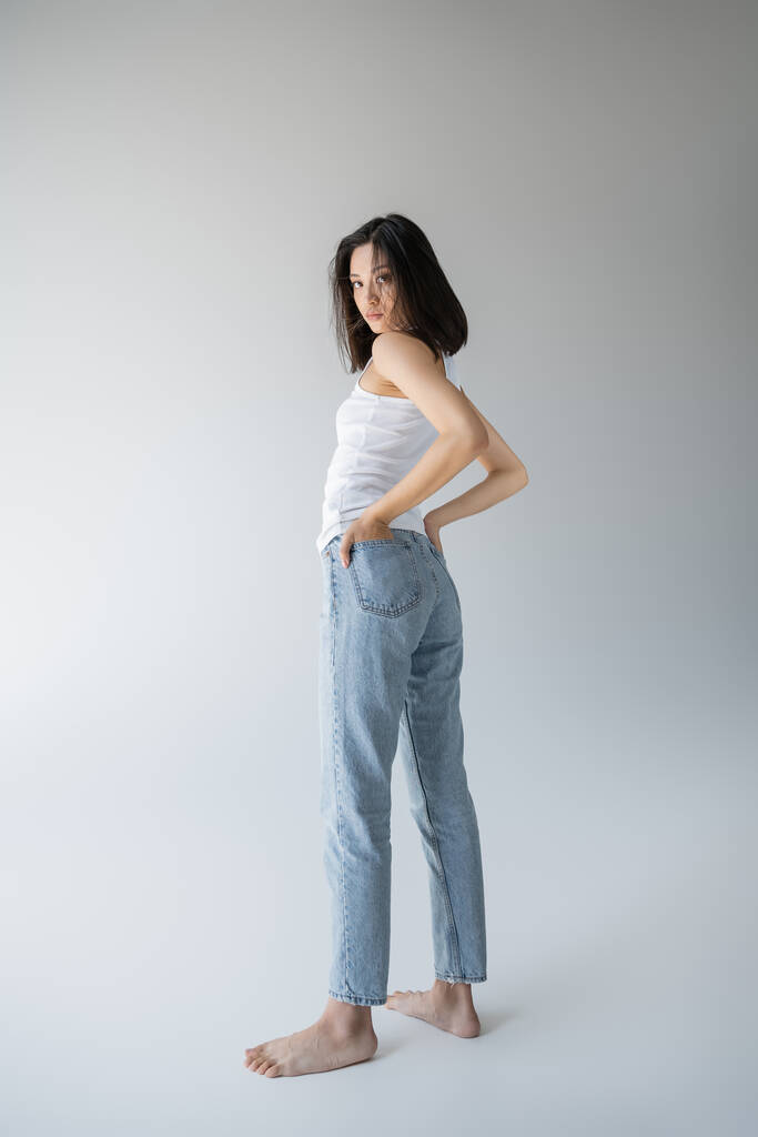 full length of barefoot asian woman in white tank top and blue jeans holding hands in back pockets on grey background - Photo, Image